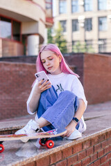 A pink-haired hipster teenage girl in a white T-shirt,jeans and with a skateboard is sitting on a...