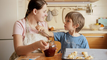 Smiling little boy feeding his mother with jam from spoon while making biscuits at home. Children cooking with parents, little chef, family having time together, domestic kitchen.