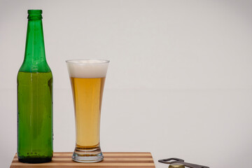 Un vaso y una botella verde con cerveza junto a la tapa de la botella y un abridor, sobre un fondo blanco, con espacio de copia