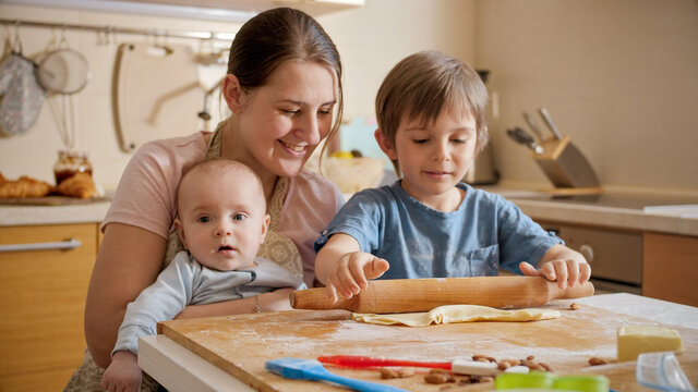 Young Mother Holding Baby Helping Older Son Baking Pizza At Home. Children Cooking With Parents, Little Chef, Family Having Time Together, Domestic Kitchen.