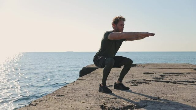 Young strong sporty athletic toned fit sportsman man 20s wear sports clothes warm up training doing legs squat at sunrise sun dawn over sea beach outdoor on concrete pier seaside in summer day morning