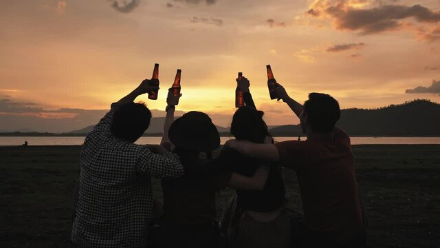 Rearview of a group of friends clinking bottles of beer together, sitting and look at the sky at sunset time. travel, camping, celebration, weekend Activities. slow motion