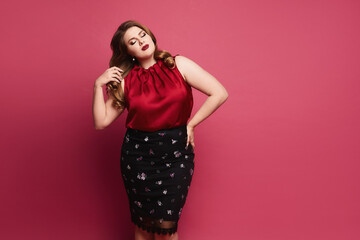 Young plus size caucasian model woman in red satin blouse and skirt posing over pink background....