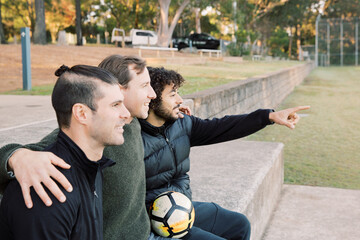 Side view photo of 3 smiling interracial man sitting on a field, one is pointing a finger