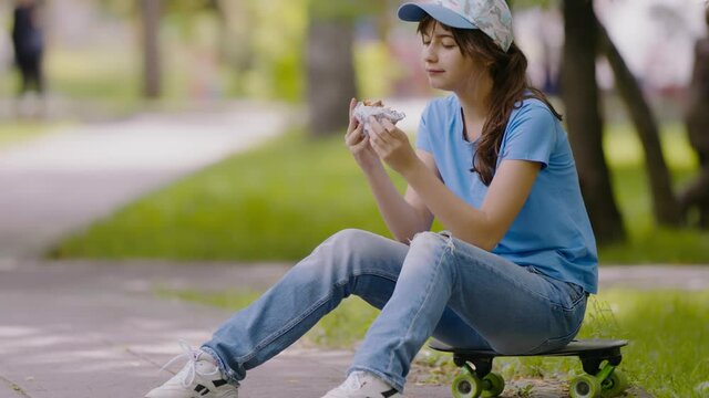Teen schoolgirl lunch outdoors. Child kid eating fast food hamburger sitting on skateboard in park