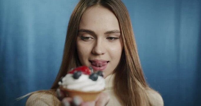 Portrait of young woman looking at delicious sweet cake with berries, licking her lips. Close-up of female student with green eyes. Happy hungry model tasting cheesecake in studio.