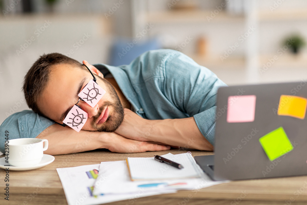 Poster lazy unproductive young guy wearing funny sticky notes with open eyes on his glasses, sleeping at wo