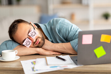 Lazy unproductive young guy wearing funny sticky notes with open eyes on his glasses, sleeping at...