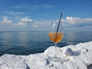 Wine glass of luxury cocktail Aperol Spritz on white stones with amazing ocean view and cloudscape. Summer vacation party drink