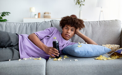 Bored black teenager lying on couch with scattered food, watching dull show or movie on TV, killing...