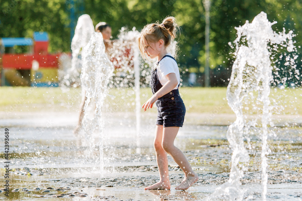 Wall mural Cute cheerful girl playing in fountain. Kid in denim overall having fun in summer park