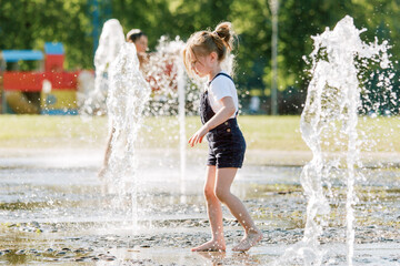 Cute cheerful girl playing in fountain. Kid in denim overall having fun in summer park