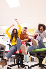 Colleagues having fun on work at the office. Girls pushing boys on chairs with wheels. Multiethnic group of successful start up company.