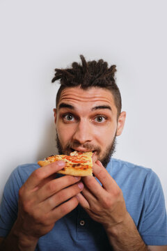 Human holds piece of cheese pizza with tomatoes in his hands and enjoys eating. Young cute Caucasian man with beard and dreadlocks eats pizza on white neutral background. Vertical image.