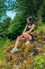 Naklejka na ściany i meble Young female athlete squats on the rocks at a small waterfall spring forest. Rest on the nature, a track, run