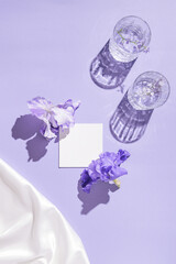 Two glasse with water and card note with iris flowers on pastel lilac background with white silk cloth. Summer refreshment concept. Sunlit flat lay. Minimal style. Top view