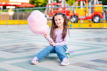 a child girl in an amusement park in the summer eats cotton candy on a skateboard and smiles with...