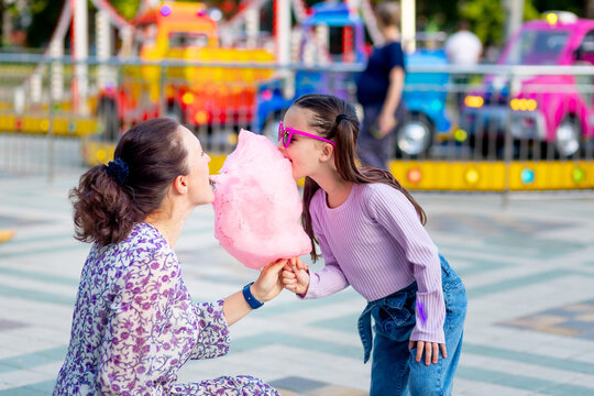 a child girl with her mother in an amusement park in the summer eating cotton candy and ice cream near the carousels, fooling around and laughing, the concept of family weekends and school holidays