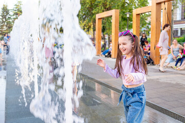 a child girl plays and bathes in the city fountain in the summer and smiles or laughs with...
