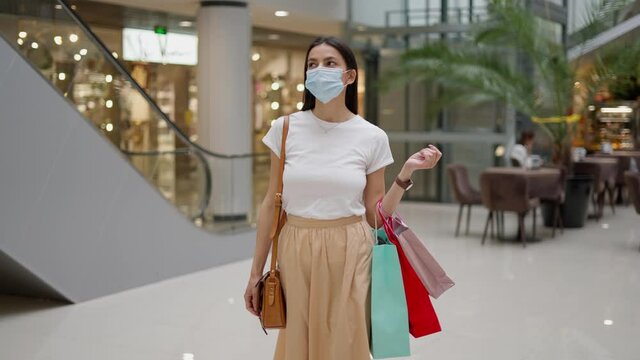 Attractive young woman in trendy clothes wearing disposal face mask carrying many paper bags, looking around mall with different fashion stores and boutiques, happy to have shopping during pandemic