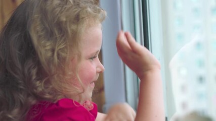 Close-up of a happy little girl seeing someone in the window. A blond, curly-haired girl waves her parents out the window and laughs.