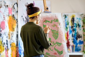Back view of young female artist creating abstract painting, standing in living room at home