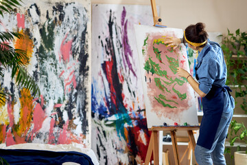 Back view of talented female artist working on a modern abstract oil painting. She is applying paint on canvas with fingers, standing in home studio workshop
