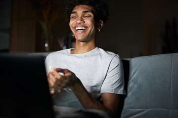 Joyful young guy laughing while watching comedy movie on a laptop, sitting on a sofa in dark room...