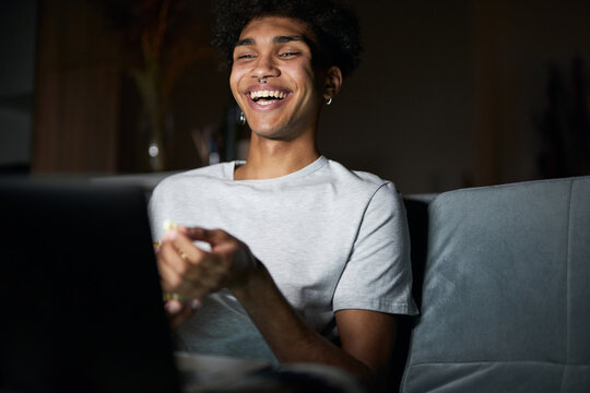 Joyful young guy laughing while watching comedy movie on a laptop, sitting on a sofa in dark room at home