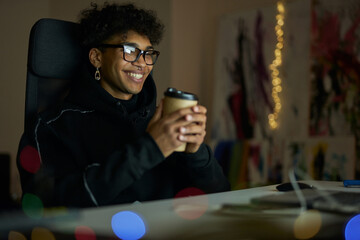 Leisure activities. Happy young guy with piercing wearing glasses holding disposable cup of coffee, sitting at the table and watching videos using laptop late at night
