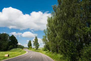 Scenic countryside landscape. Empty asphalt road. Summer nature. Blue cloudy sky. Green trees. Haystack on grass.