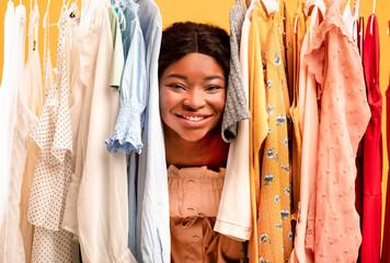 Young black woman peeking out between clothes hanging in rail, smiling at camera, satisfied with shopping choice