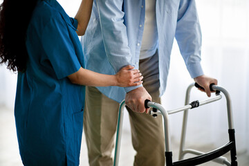 Cropped view of elderly man using walker, young nurse supporting and assisting him at retirement home, closeup