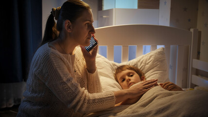 Young worried mother writing to doctor on smartphone next to sick little son lying in bed. Concept...