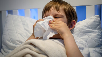 Portrait of little sick boy with runny nose lying in bed and blowing nose in paper tissue. Concept...