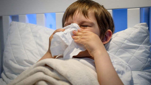 Portrait Of Little Sick Boy With Runny Nose Lying In Bed And Blowing Nose In Paper Tissue. Concept Of Children Illness, Disease And Healthcare.