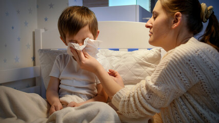Little sick boy blowing nose in paper tissue and talking to mother before going to sleep at night. Concept of children illness, disease and parent care