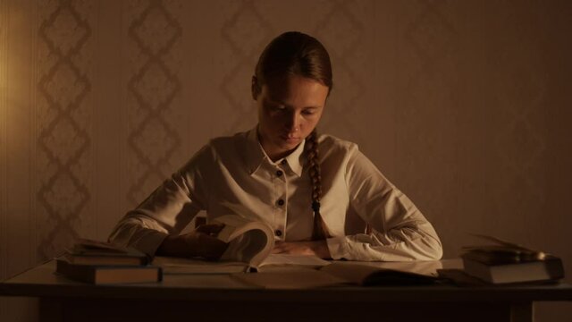 Young woman sitting at the table with books in the evening and flipping pages of  encyclopedia book. Education, studying at home, library, doing homework, prepare for examination. Student lifestyle.
