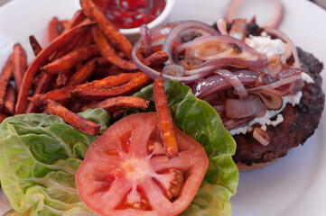 Image of a hamburger with sweet potatoes fries, tomato slice, lettuce, and onions on a white dish.