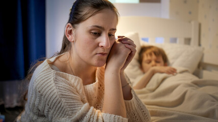 Young mother sitting next to bed of her sleeping son suffering from depression and bad thoughts....