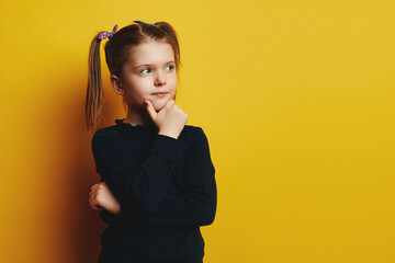 Smart wunderkind kid girl touching cheek and looking up while thinking against blue background 