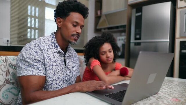 Black father and daughter in front of laptop computer together at home kitchen table