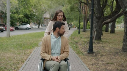 Slow motion of caring young lady pushing man's wheelchair talking smiling outdoors in park. Assistance for disabled people and lifestyle concept.