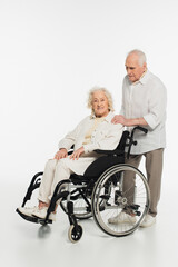 Fototapeta na wymiar elderly man holding hand on shoulder of wife in wheelchair isolated on white