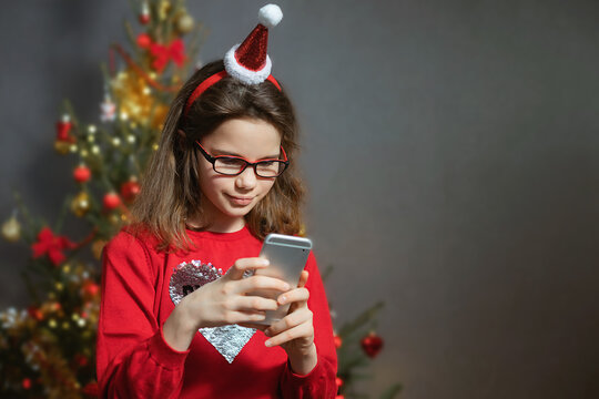 A cute girl  in  glasses in a red sweater and a stanta claus hat with a smartphone in his hands is typing a message against the Christmas tree indoors. Copy space.