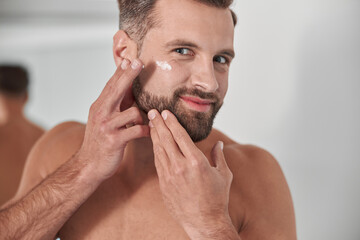 Happy man with stylish beard applies antiaging cream onto crowl feet on face in bathroom
