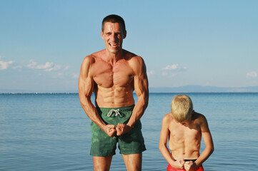 Father and son having fun on the beach and showing muscles . Being a good example to your child concept - obrazy, fototapety, plakaty
