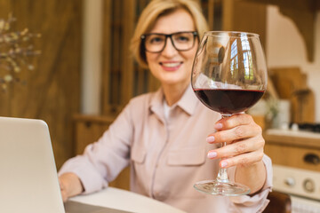 Portrait of smiling mature senior woman holding glass of wine while using laptop on kitchen table. Freelance working at home concept.