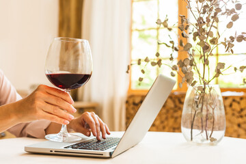 Portrait of smiling mature senior woman holding glass of wine while using laptop on kitchen table. Freelance working at home concept.