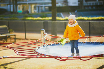 Adorable toddler girl in yellow jacket having fun on a trampoline on a fall or spring day